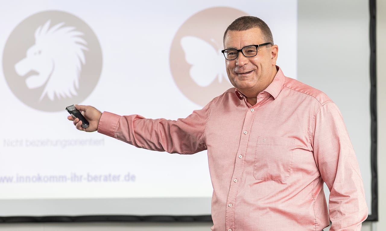 Alexander Preidt im roséfarbenen Hemd zeigt mit dem Presenter auf eine Präsentationsfolie